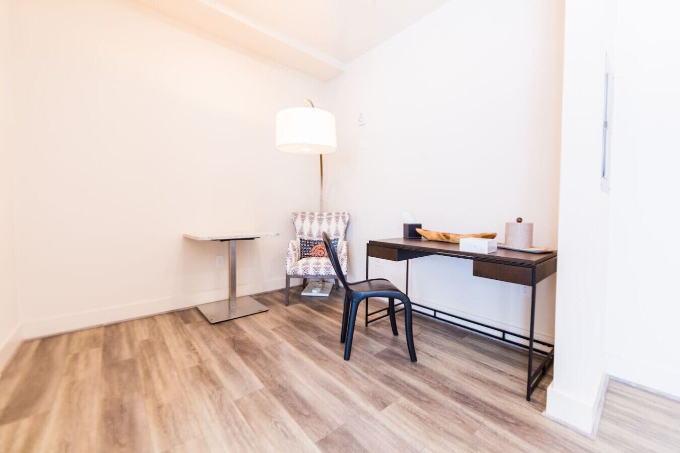 A small, well-lit room with a wooden floor, a desk with a chair, a lamp, and a small table.