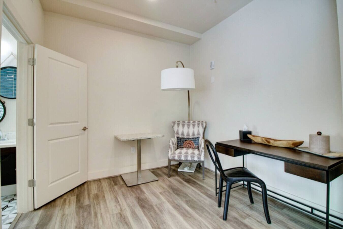 A small, compact room with a wooden floor, a white door, a small table with a chair, a lamp, and a wall-mounted shelf.