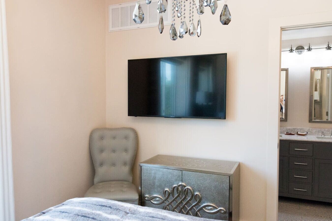A bedroom with a bed, a chair, a dresser, a TV mounted on the wall, and a bathroom with a mirror and sink.