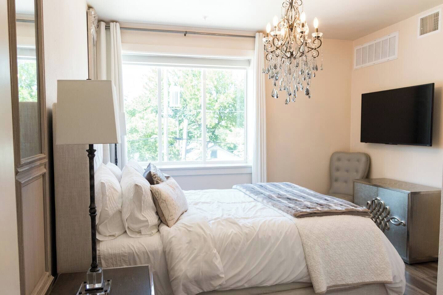 A bedroom with a large bed, a chandelier, a television, and a window with a view of trees.