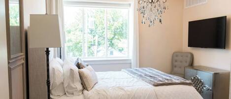 A bedroom with a large bed, a chandelier, a television, and a window with a view of trees.