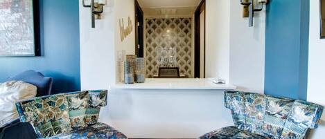 Quarto com duas cadeiras estampadas, uma mesa pequena e uma parede decorativa com um letreiro de 'Welcome'.