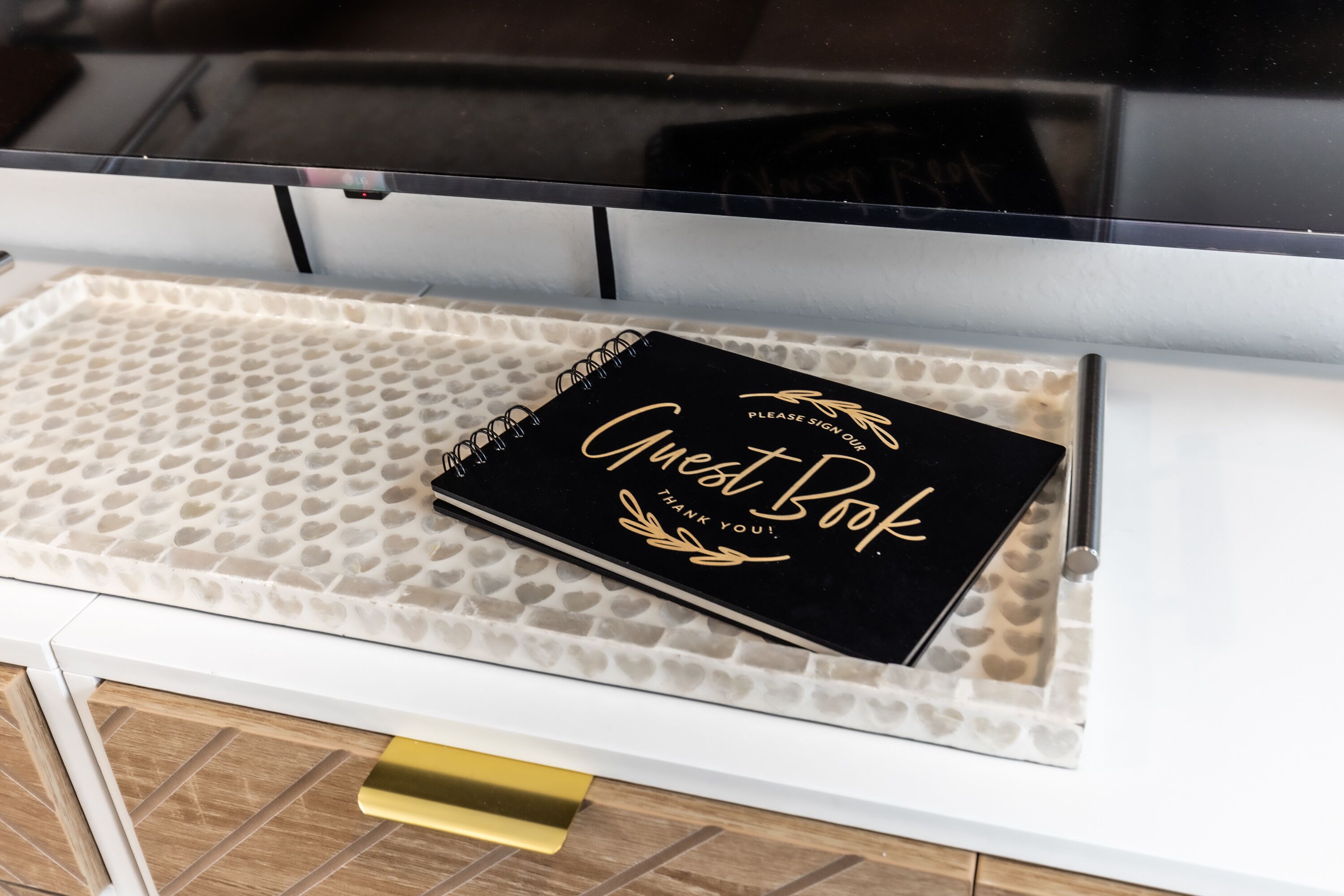 A black guest book on a white tray with a heart-patterned mat, placed on a white cabinet with a gold handle.
