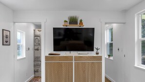 Una sala moderna con un televisor de pantalla plana montado en un mueble de madera, un estante pequeño con plantas y una puerta que da a otra habitación.