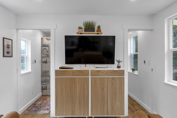 Una sala moderna con un televisor de pantalla plana montado en un mueble de madera, un estante pequeño con plantas y una puerta que da a otra habitación.