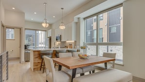 Une cuisine moderne dotée d’un îlot central, d’un coin repas avec une table et des chaises en bois, et de grandes fenêtres offrant une vue sur la ville.
