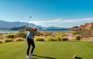 Une personne jouant au golf sur un parcours bien entretenu, avec une vue pittoresque sur les montagnes et l’océan en arrière-plan.