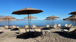 Une plage avec des chaises longues et des parasols, une mer d’un bleu limpide et des montagnes à l’horizon.