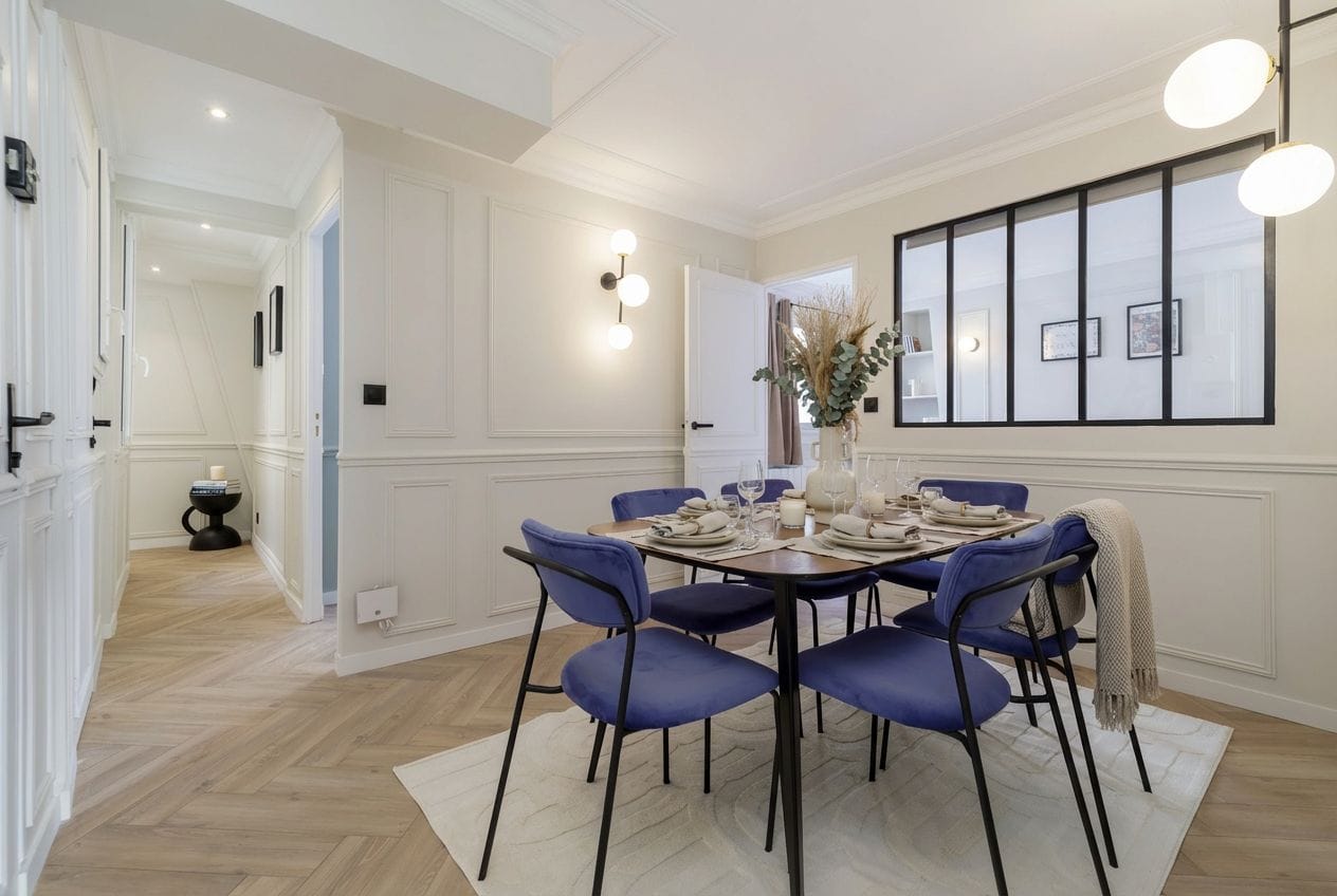 Un comedor con una mesa redonda dispuesta para cuatro personas, sillas tapizadas en azul, un florero con plantas secas y una lámpara en la pared.