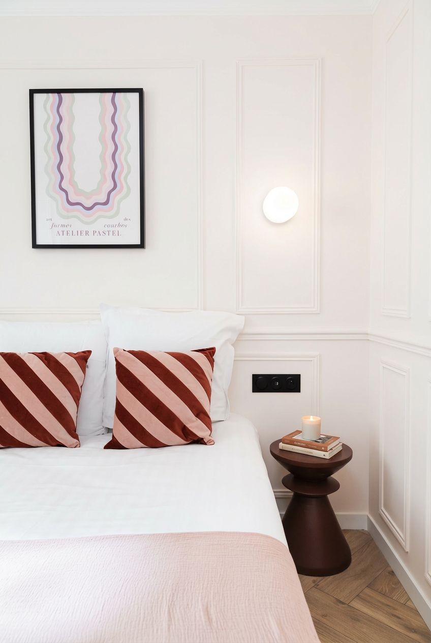 Una cama bien hecha con almohadas a rayas, una obra de arte enmarcada encima y una mesita de noche con un libro y una vela.