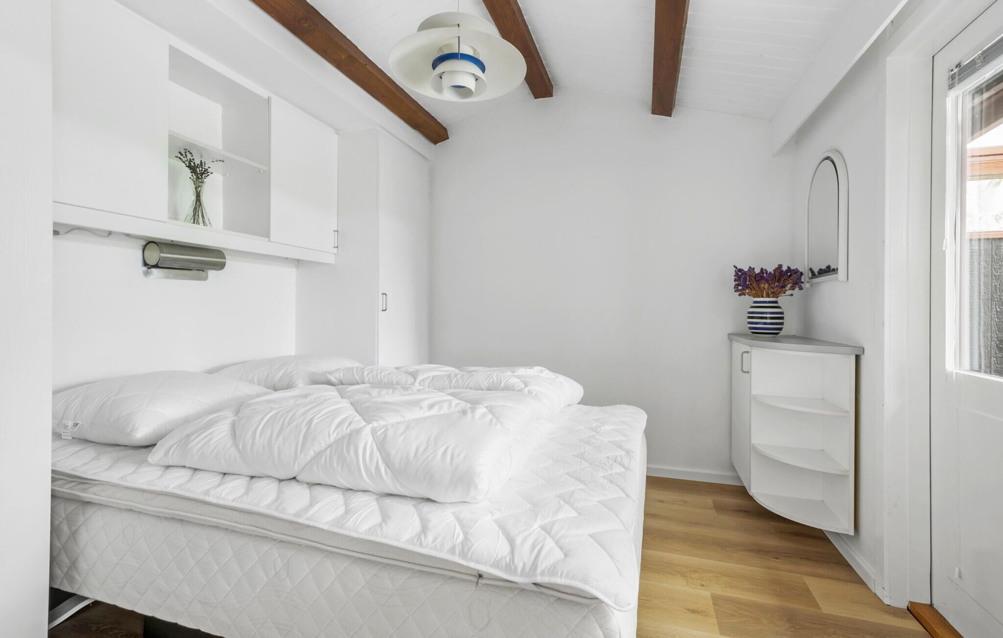 Une chambre à coucher avec un lit, une table de chevet ornée d’un vase, un miroir et une étagère sur laquelle sont disposés des objets décoratifs.