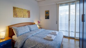 Ein Hotelzimmer mit Bett, Nachttisch, Lampe, Fenster mit Blick und Kunstwerk an der Wand.