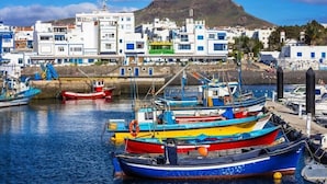A harbor with colorful boats docked, a mountainous backdrop, and buildings with blue and white facades. - Paraisos de Agaete Penthouse Views (El Turman)