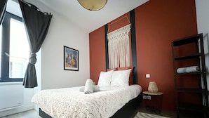 Une chambre à coucher avec un lit, un tableau, une décoration murale en macramé, une lampe et une étagère avec des serviettes.