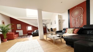 Un salon spacieux avec un grand canapé en cuir noir, une table basse en verre et un tapis blanc. Un mur rouge est décoré d’un attrape-rêves, et le sol est en bois.