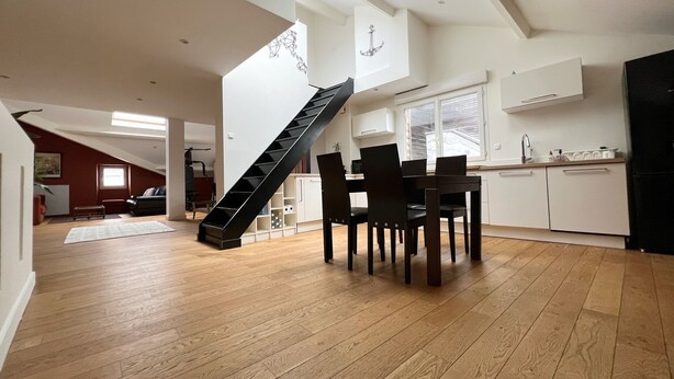 Un vaste loft doté d’un plancher en bois, d’un coin repas avec une table et des chaises, d’une cuisine avec des meubles blancs et d’une lucarne.
