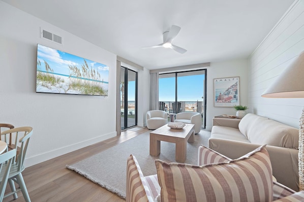 A modern living room with a flat-screen TV, a ceiling fan, a sofa, a coffee table, and a dining area with chairs.