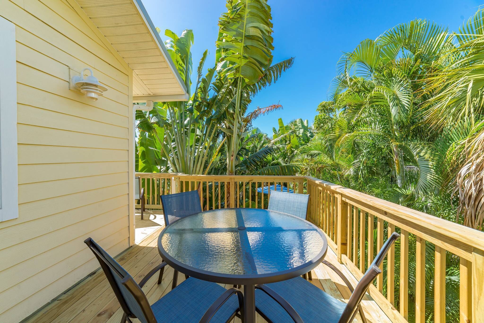 A wooden deck with a round glass table and chairs, surrounded by lush tropical plants.