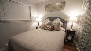 Une chambre à coucher avec un grand lit, deux tables de chevet avec des lampes, une tête de lit agrémentée de coussins décoratifs et un mur orné d’une œuvre d’art texturée.
