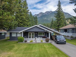 A single-story house with a dark exterior, white porch, and a garage. It has a number '63' and '7320' on the front, a small front yard, and a mountain backdrop.