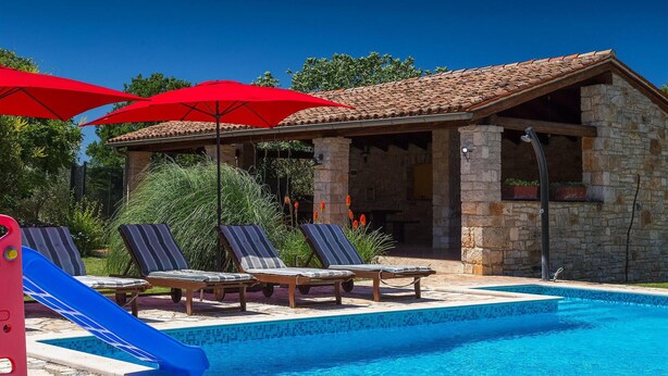 A pool area with lounge chairs and a slide, a stone building with a tiled roof, and red umbrellas. - Beach front home in Pula (Pula)