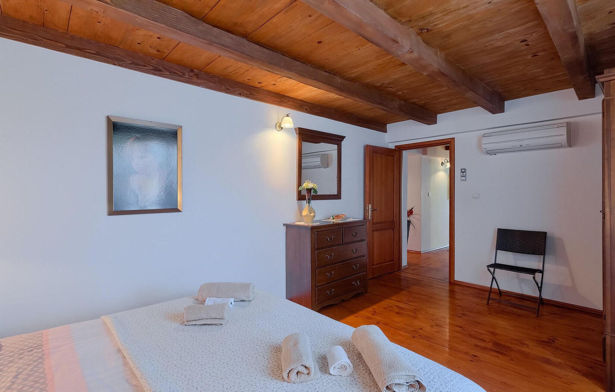 Ein Schlafzimmer mit Bett, Kommode, Spiegel und Stuhl. Eine Tür führt in einen anderen Raum.