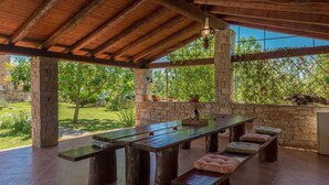 An outdoor patio with a long wooden table and benches, stone walls, and a view of greenery. - Beach front home in Pula (Pula)