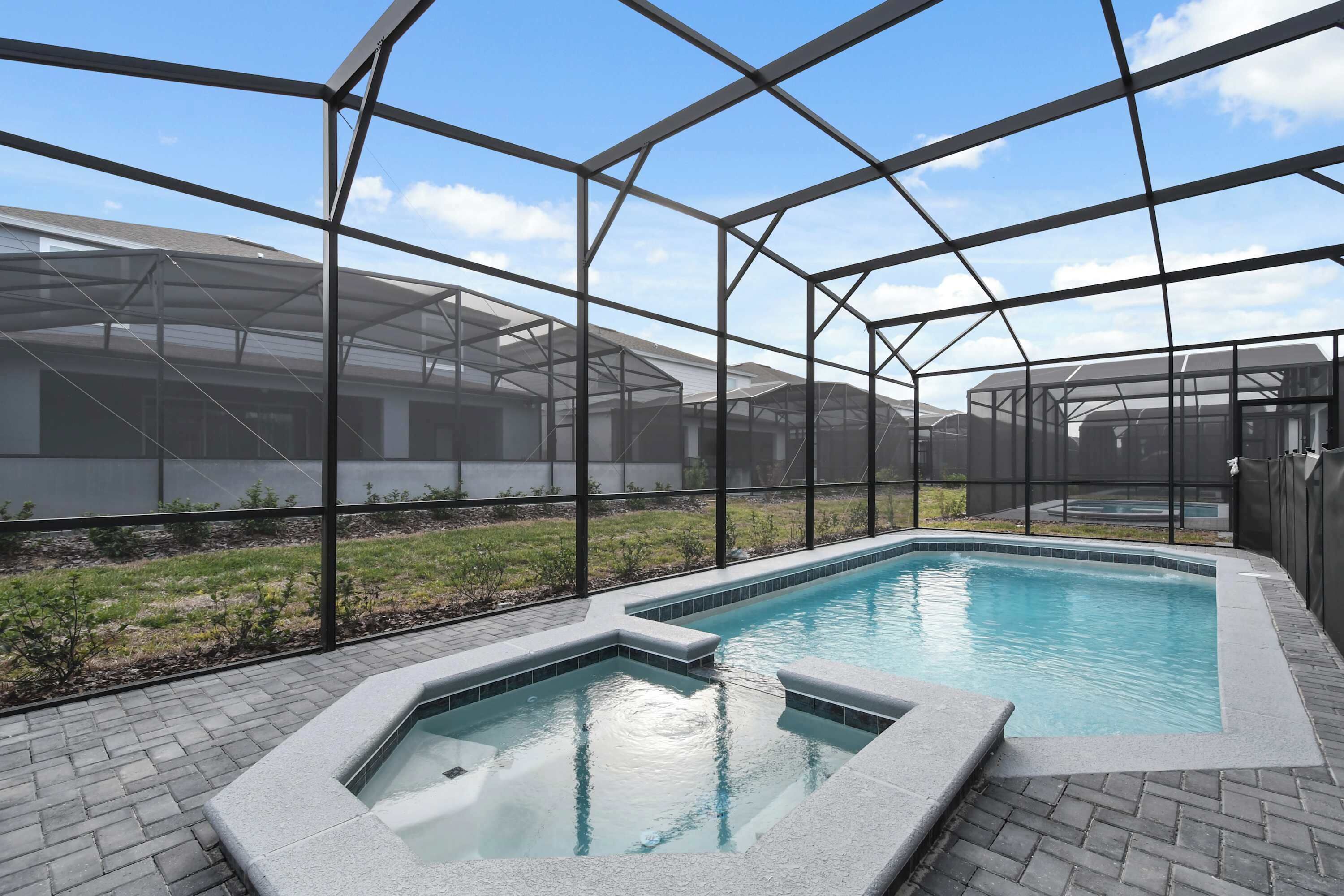 Una piscina con cerramiento de cristal, rodeada por un patio de ladrillo y una zona ajardinada.