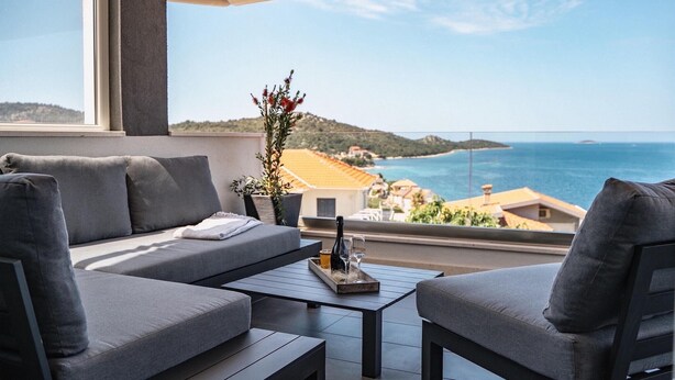A balcony with a gray sofa, two armchairs, and a coffee table. A potted plant with red flowers is on the table. The view includes a coastal landscape with buildings and a clear sky. - Pet friendly home in Razanj (Razanj)