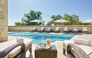 Un espace aménagé au bord de la piscine, avec des chaises longues et des parasols, une petite table avec des boissons et des verres, et un mur de pierre en arrière-plan.