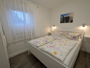 Makuuhuone, jossa on sänky, valkoiset verhot, yöpöytä, seinään kiinnitetty lamppu ja kehystetty kuva.