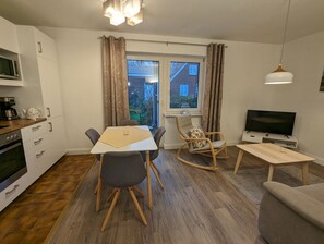 Moderni keittiö, jossa on mikroaaltouuni, liesi ja kahvinkeitin. Ruokailutilassa on pöytä ja tuolit, keinutuoli ja televisio. Huoneessa on suuret ikkunat, joissa on verhot.