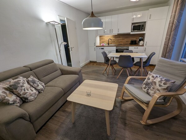 Un salon moderne avec un canapé, une table basse et un fauteuil à bascule. Il y a également un coin cuisine avec des meubles blancs et un four à micro-ondes.