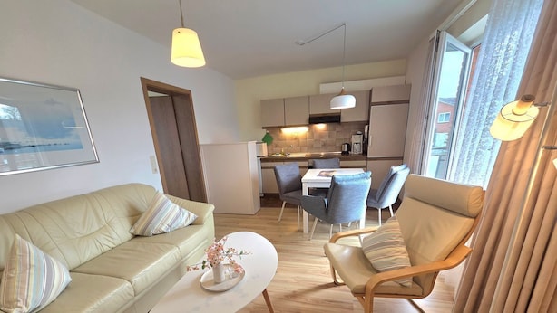 Una sala moderna con sofá, sillón y mesa de centro, un comedor con sillas y una cocina al fondo.
