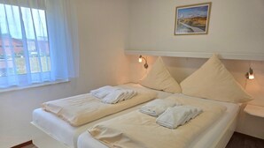 Cama doble con dos almohadas y toallas dobladas, mesita de noche con lámpara, y ventana con cortinas traslúcidas.