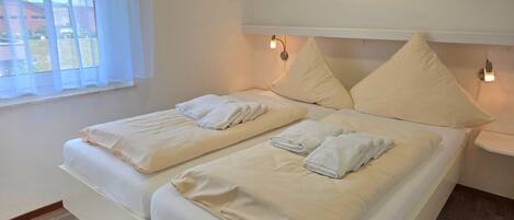 Cama doble con dos almohadas y toallas dobladas, mesita de noche con lámpara, y ventana con cortinas traslúcidas.