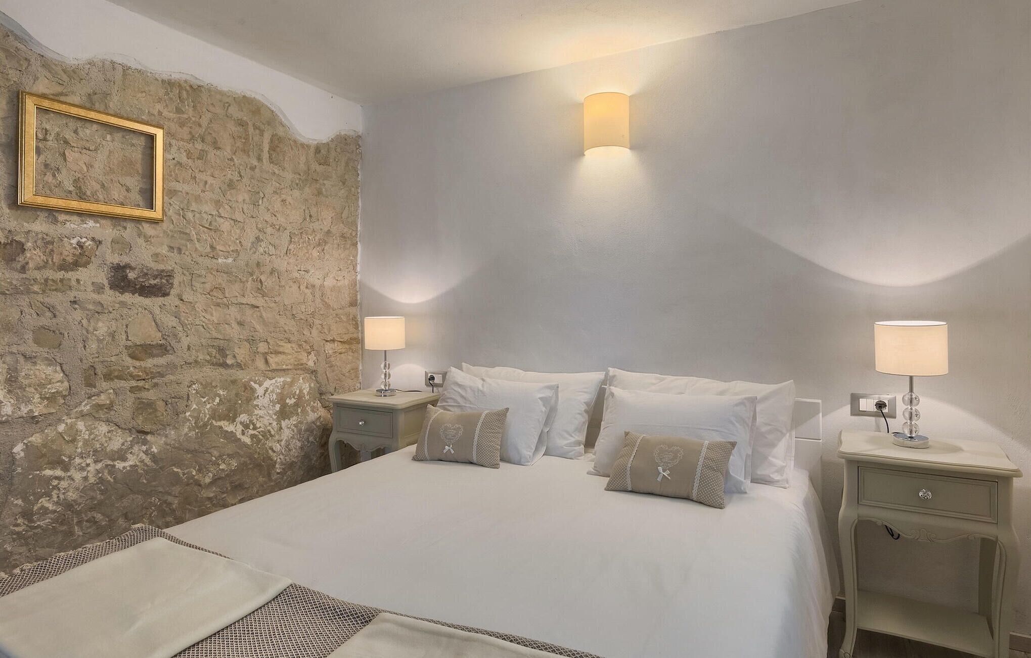 A bedroom with a stone wall, a bed with white bedding, two bedside tables with lamps, and a small framed picture on the wall.