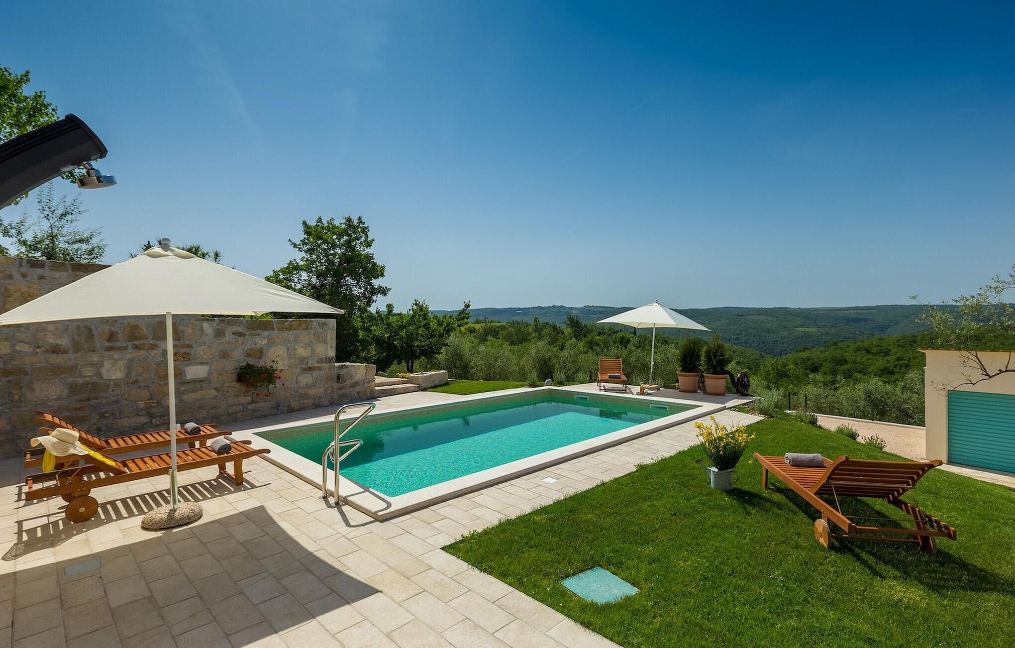 A swimming pool with a stone wall and a grassy area with sun loungers and umbrellas.