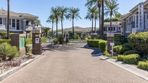 Una entrada protegida por un portón a un complejo residencial, con palmeras y setos bien cuidados.