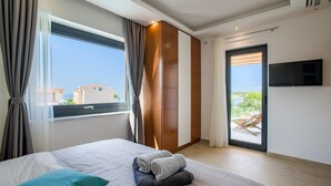 A modern hotel room with a large bed, a view of the sea, a sliding door to a balcony, and a flat-screen TV mounted on the wall.