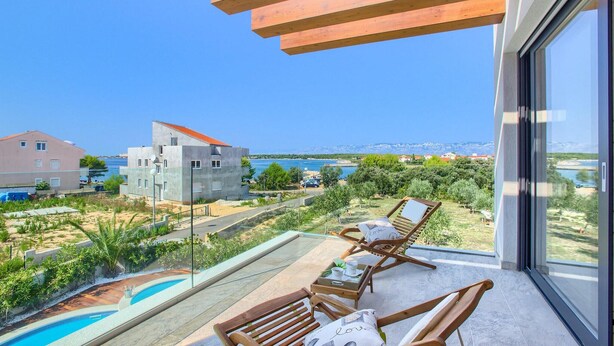 A balcony with a pool, lounge chairs, and a view of the sea and buildings.