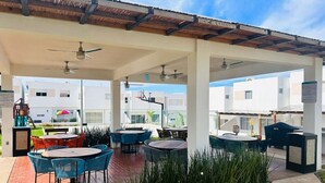 An outdoor patio area with multiple tables and chairs, covered by a roof with ceiling fans, and surrounded by buildings.