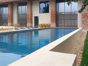 A rectangular swimming pool with a tiled deck, surrounded by brick walls and large windows. - We Rural by Interhome (Poirino)