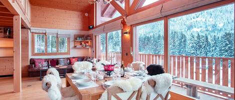 A wooden cabin interior with a dining table set for six, large windows offering a view of snow-covered trees, and a chandelier with antler-shaped light fixtures.