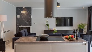A modern kitchen with a marble countertop, a built-in stove, and a large black range hood. There is a seating area with a sofa and a dining table with a bowl of fruit.
