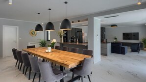 A modern dining area with a wooden table and grey upholstered chairs, a kitchen in the background, and pendant lighting.