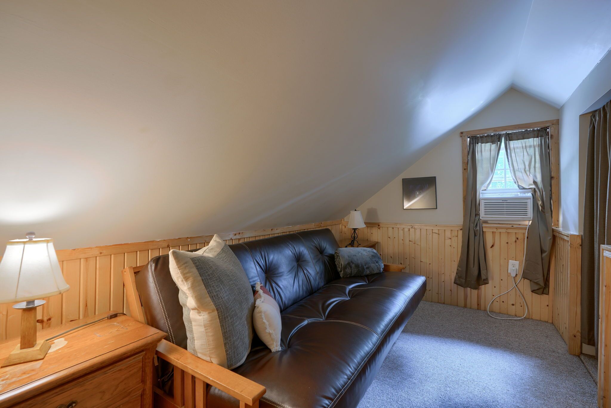 Una habitación acogedora con un sofá de cuero, una mesita de noche de madera, una lámpara y una ventana con cortinas.