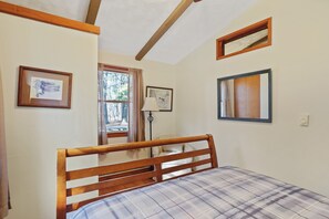 A bedroom with a wooden bed frame, a plaid bedspread, a window with curtains, and two framed pictures on the wall.