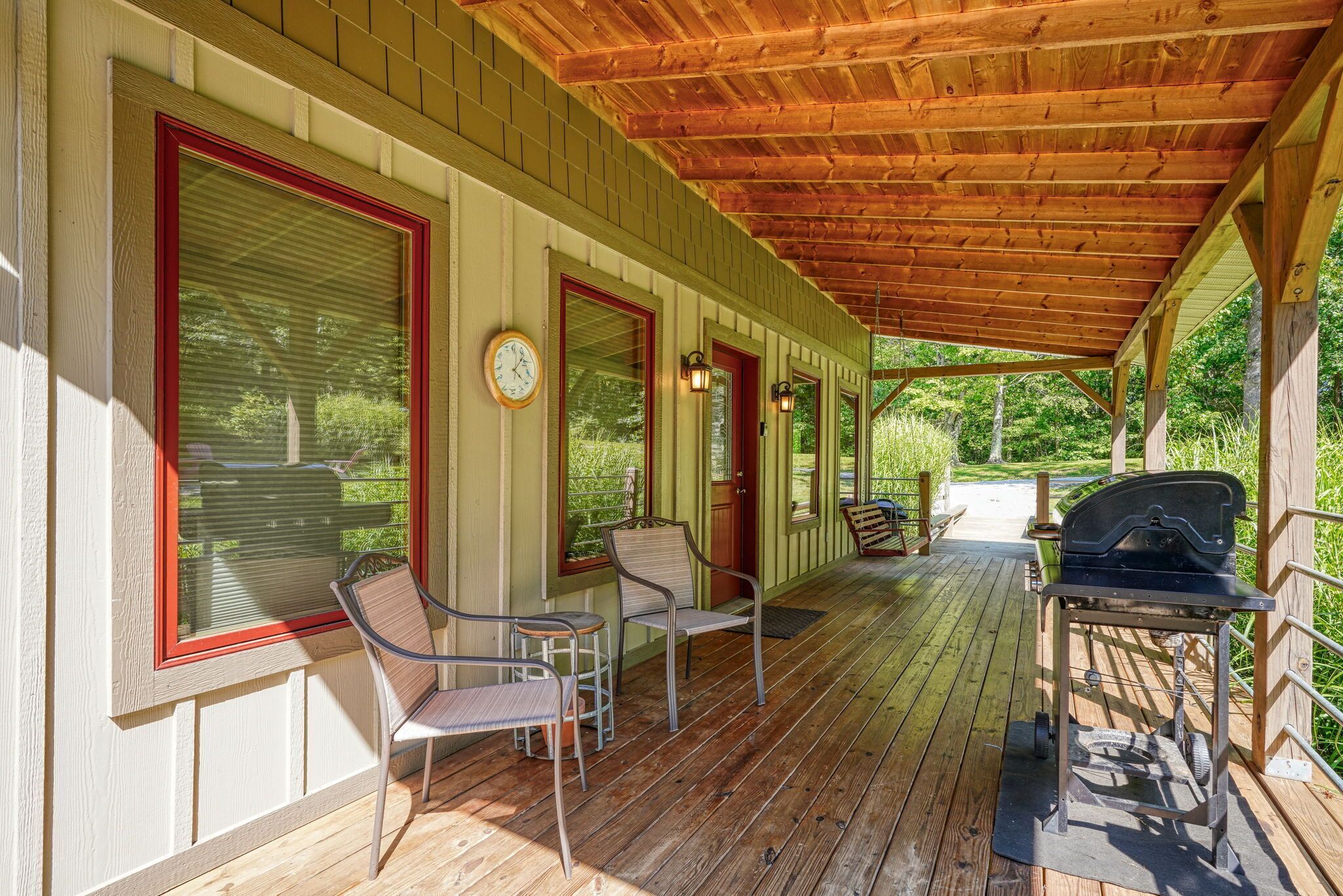 A covered porch with wooden flooring, metal chairs, a barbecue grill, and a clock on the wall.