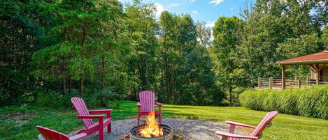 A fire pit with four Adirondack chairs arranged around it, set on a gravel area with a grassy lawn and trees in the background.